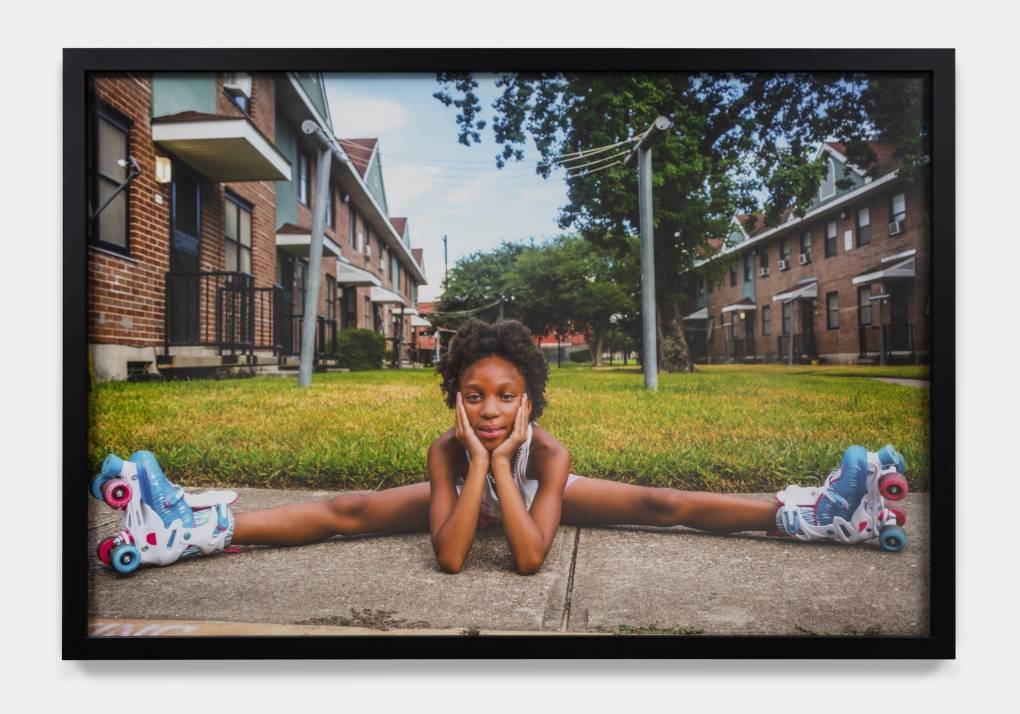 A little girl sits on the sidewalk in the splits while wearing roller skates.