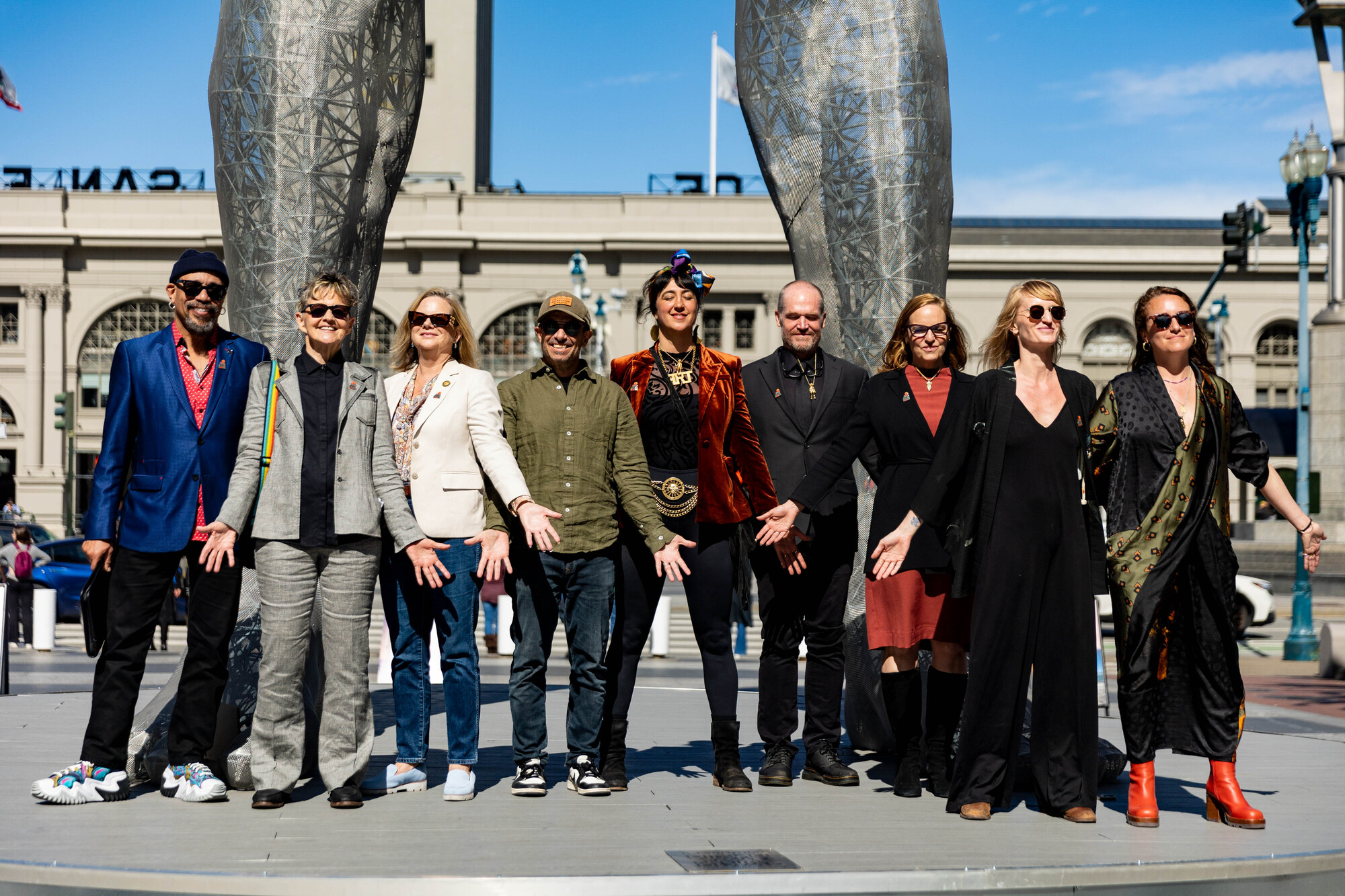 nine people stand on platform in mountain yoga pose with eyes closed, giant sculpture legs behind them