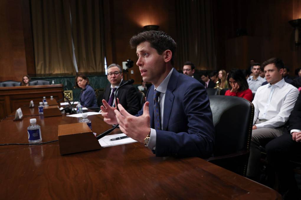 A white man in a blue suit and tie gestures as he speaks in a congressional room surrounded by people.