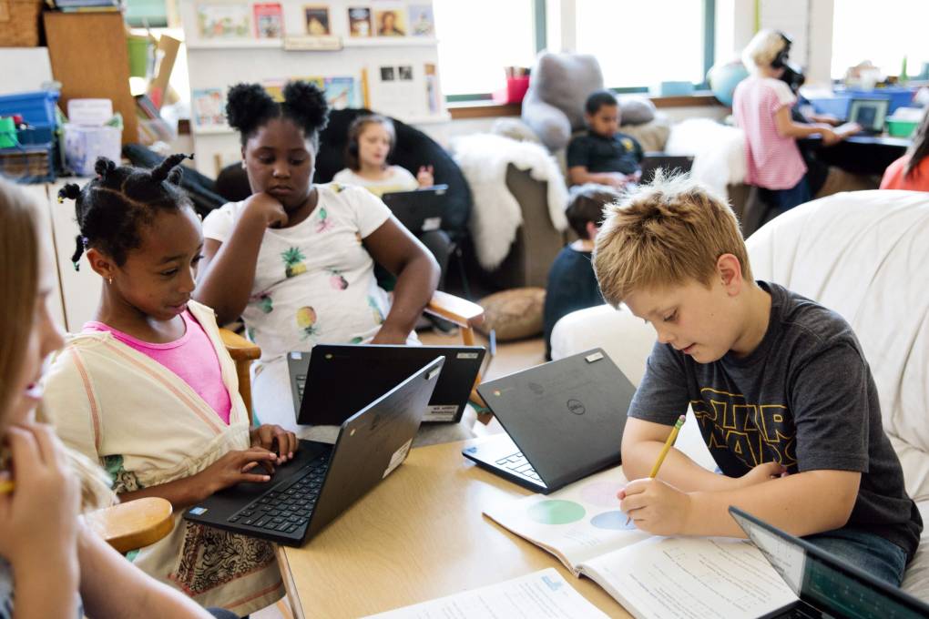 Students in a classroom on computers or writing in notebooks. Sunlight streams in through the windows, the children look happy.